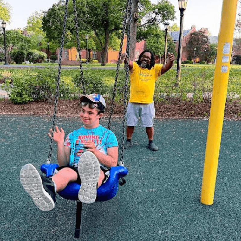 A smiling teacher pushes a happy student on an accessible swing