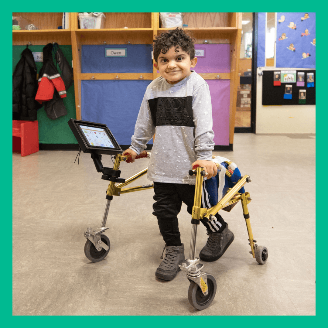 Amir stands with a gait trainer in a classroom.