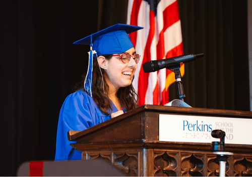 Sophia stands at a podium in a cape and gown, speaking into a microphone.