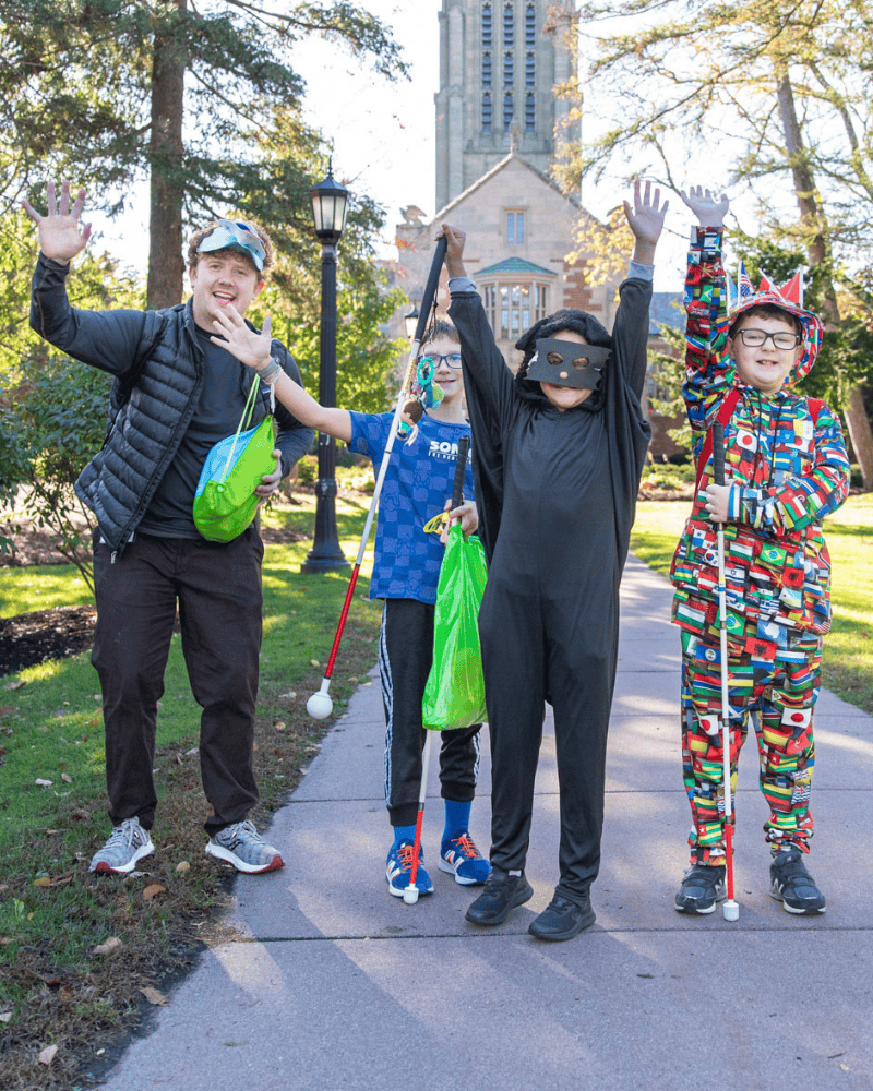 An adult and three children wearing Halloween costumes raise their hands in the air and smile. Two of the children are holding white canes.